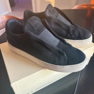 Svarta sneakers från Axel Arigato - Stilrena svarta sneakers från Axel Arigato i mocka med vit platt sula. Skorna har minimalistisk design utan snörning och diskret logga på sidan. Kommer med originalkartong och dustbag. Perfekta för dig som gillar clean och modern look.