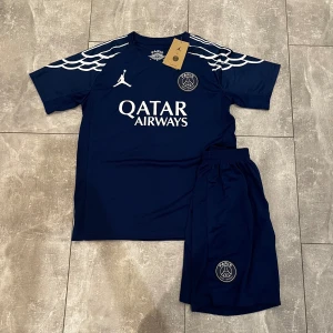 PSG marinblå set Jordan - Marinblå PSG set från Jordan med vita detaljer och tryck. Kortärmad modell med vågigt mönster på axlarna, Qatar Airways-logga och PSG-emblem på bröstet. Tillverkad i lätt, ventilerande material som passar perfekt för träning.