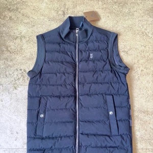 Svart quiltad dunväst från Burberry - Snygg svart dunväst från Burberry med quiltad framsida, dragkedja och två fickor med knapp. Baksidan har klassiskt rutig Burberry-mönster i blå och grå toner. Perfekt för lager-på-lager och har en stilren ståkrage.