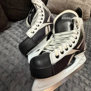 Svarta och vita hockeyskridskor Reebok 41 - Grymma hockeyskridskor från Reebok i storlek 41. De är svarta och vita med klassisk Reebok-logga, snörning och tåskydd i plast. Yttermaterialet är syntet och insidan är fodrad för extra komfort. Perfekta för isen och redo för nästa match!