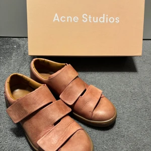  Acne Studios - Unika rosa sneakers från Acne Studios i skinn med två breda kardborreband över foten. Skorna har en rund tå och beige innersula, samt en diskret gummisula. Perfekta för dig som gillar stilrena och trendiga detaljer. Acne Studios triple Lo. Sparsamt använda. Storlek 37.