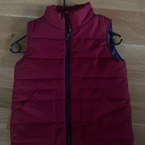 Röd dunväst från H&M med fickor - Snygg röd dunväst från H&M med hög krage och två stora fickor framtill. Västen har en mörk dragkedja och quiltad design som ger en cool look. Perfekt att slänga över en hoodie när det är lite kyligt ute.