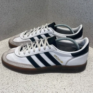 Adidas Spezial vita och svarta sneakers - Adidas Spezial sneakers i vitt och svart med klassiska tre ränder på sidan, gummisula i brunt och detaljer i grå mocka framtill. Skon har snörning och rund tå, samt guldfärgad Spezial-logga på sidan.