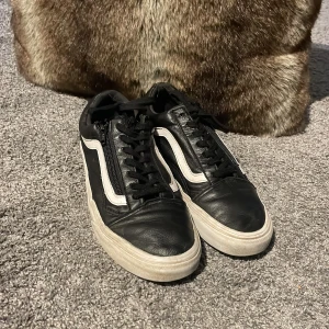 Svarta Vans med dragkedja och läder skinn  - Klassiska Vans Old Skool sneakers i svart skinn med vit sula och den ikoniska vita sidoremsan. Skorna har snörning och låg profil, samt den typiska gummisulan med rutmönster. Perfekta för en streetwear-look och funkar året runt.