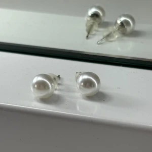 Pärlörhängen i silverfärg - Säljer ett par klassiska pärlörhängen med silverfärgad infattning. Örhängena har rund form och en elegant, vit pärlyta som ger en tidlös och stilren look. Perfekta för dig som gillar enkel men snygg design.