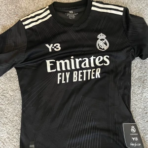 Real Madrid Y-3 svart matchtröja !skadad på ryggen! - Snygg svart Real Madrid fotbollströja från Adidas och Y-3 med vita detaljer och klubbmärke på bröstet. Tröjan har korta ärmar, klassiska tre ränder på axlarna och ett subtilt grafiskt mönster över hela tyget. Materialet är lätt och andas, perfekt för träning eller match.