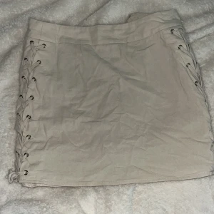 Beige kortkjol med snörning - Säljer en beige kortkjol med snygg snörningsdetalj längs ena sidan. Kjolen har en tight passform och sitter väldigt fint och perfekt. Gjord för sig som vill ha en kjol som sitter tight och fint!💕 Nästan aldrig använd och väldigt fint skick❣️