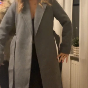 Lång Grå kappa med bälte och bred krage - Snygg grå kappa med bred krage och knytskärp i midjan. Lång modell med raka ärmar och stora fickor framtill. Perfekt för dig som vill ha en stilren och klassisk look under kyliga dagar. Materialet känns mjukt och värmande. Sällan använd som ny! 