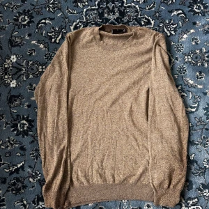 Beige rundhalsad tröja Oscar Jacobson - Snygg beige rundhalsad tröja från Oscar Jacobson, tillverkad i Italien. Tröjan är stickad och har långa ärmar med ribbade muddar vid hals, ärmslut och nederkant. Perfekt för dig som gillar klassisk och stilren design.