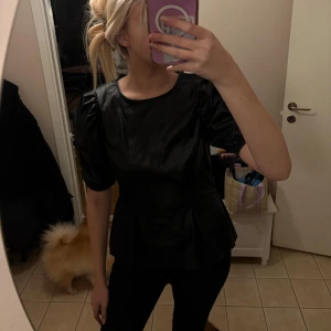 Svart puffärmad topp i skinnimitation - Svart topp från Gina Tricot i skinnimitation med rund halsringning och korta puffärmar. Snygg figurnära passform med lätt utsvängd nederdel. Perfekt för dig som vill sticka ut med en edgy look.