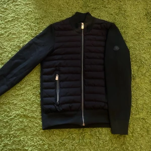Moncler cardigan - Svart pufferjacka från Moncler med quiltad front och släta ärmar. Jackan har två dragkedjeförsedda fickor, ribbad krage och Moncler-logga på ärmen. Materialet är en mix av polyester och bomull, perfekt för höst och vinter. Snygg och stilren design med normal passform.