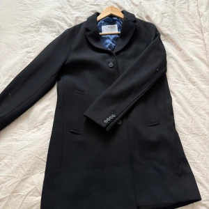 Svart rock från Selected Homme - Stilren svart rock från Selected Homme med klassisk krage, knäppning framtill och diskreta fickor. Jackan har helfodrad insida i blått och grått tyg och fyra knappar vid ärmslut. Perfekt för dig som gillar en clean och tidlös look.