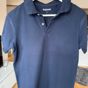 Marinblå pikétröja från Kappahl - Snygg marinblå pikétröja från Kappahl i storlek S. Klassisk krage, korta ärmar och två knappar framtill. Tröjan är gjord i mjuk bomull och har en stilren look som funkar till många olika outfits.