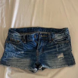 Blå jeansshorts från FB Sister - Säljer ett par blå jeansshorts från FB Sister med coola slitningar och fransig kant. Shortsen har klassisk femficksdesign, låg midja och detaljer som dragkedja och knapp framtill. Baksidan har ett brunt lädermärke med döskalle. Perfekta för sommaren!