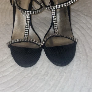 Svarta  klack - Snygga svarta sandaler med smal klack och öppen tå. Remmarna är dekorerade med glittrande strass och skon har en lätt ormskinnsstruktur. Perfekta för att lyfta din outfit med lite extra bling.