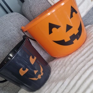 Halloween hinkar - Två Halloween godishinkar i plast, en orange och en svart, båda med pumpaansikten. Perfekt för trick-or-treat eller dekoration. Handtag i plast, gott skick utan synliga sprickor eller större slitage mer än att lite små revor på pumpaansikterna. Rymmer mycket godis och är lätta att bära.