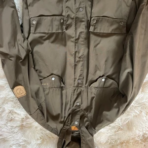 Grön Fjällräven vindjacka M - Säljer en grön vindjacka från Fjällräven i klassisk modell med fyra fickor framtill och tryckknappar. Jackan har en snygg patch på ärmen och är tillverkad i slitstark bomull/polyester-blandning. Perfekt för dig som gillar outdoor och streetstyle.