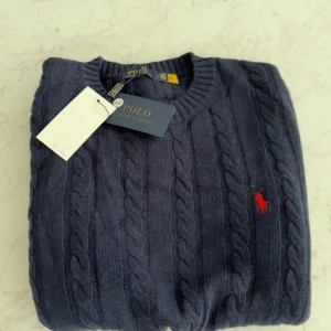 Mörkblå kabelstickad tröja Polo Ralph Lauren - Kabelstickad mörkblå tröja från Polo Ralph Lauren med klassisk röd broderad logga på bröstet. Tröjan har rund halsringning och långa ärmar, perfekt för lager-på-lager. Tillverkad i mjuk bomull för en skön känsla.