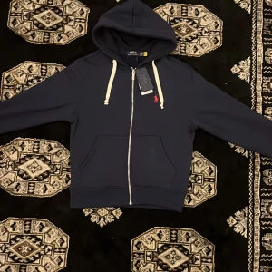 Marinblå hoodie från Polo Ralph Lauren - Marinblå hoodie från Polo Ralph Lauren med vit snörning, dragkedja och klassisk röd broderad logga på bröstet. Tröjan har huva, känguruficka och är tillverkad i mjuk bomullsmix. Perfekt för chill dagar och snygg streetstyle.