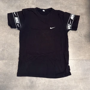 Svart Nike t-shirt med logga - Svart t-shirt från Nike i 100% bomull. Klassisk rund hals och korta ärmar med breda svarta band där Nike-loggan och swoosh syns i vitt. Swoosh-logga även på bröstet. Perfekt för dig som gillar sportig och clean streetstyle.