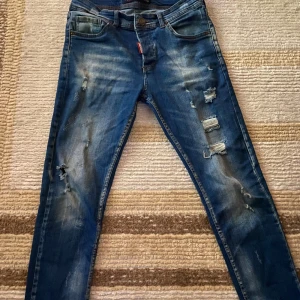 Blå Dsquared2 ICON jeans med slitningar - Säljer ett par blå Dsquared2 ICON jeans med coola slitningar och distressed detaljer framtill. Klassisk femficksmodell med bruna och röda logotypdetaljer. Jeansen har en smal passform och är tillverkade i jeansmaterial med snygg tvätt.