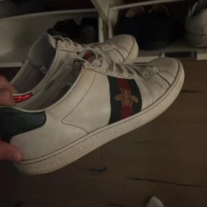 Gucci Ace sneakers med bi-detalj (1 tidigare ägare” - Gucci Ace sneakers i vitt skinn med klassiska gröna och röda ränder på sidan och en broderad guldfärgad bi. Baksidan har metallicröd och metallicgrön häl med Gucci-logga. Snygg och ikonisk modell med snörning och platt sula.