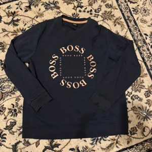 Mörkblå sweatshirt från Hugo Boss - Snygg mörkblå sweatshirt från Hugo Boss med stort BOSS-tryck i ljusrosa på bröstet. Tröjan har rund hals, långa ärmar och ribbade muddar. Perfekt för dig som gillar stilrena och trendiga märkesplagg med en clean look.