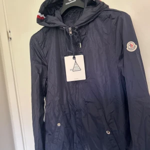 Marinblå vindjacka från Moncler - Snygg marinblå vindjacka från Moncler med huva och dragkedja framtill. Jackan har Moncler-logga på ärmen och coola rödvita detaljer på huvan. Lätt och smidig design i syntetmaterial, perfekt för blåsiga dagar. Fickor med tryckknappar och justerbar huva.