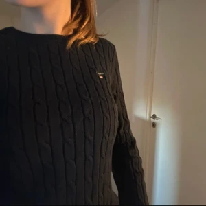 Tröja  - Svart stickad  tröja från GANT med rund hals och diskret logga på bröstet. Tröjan är gjord i stretchig bomull och har långa ärmar samt ribbade muddar vid ärmslut och nederkant. Perfekt för dig som gillar klassisk och stilren look.