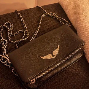 Zadig axelväska med kedjerem - Snygg svart axelväska i skinn med silverfärgad kedjerem och ett coolt emblem med vingar framtill. 