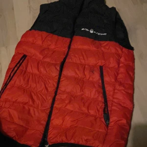 Röd och svart dunväst från Sail Racing  - Säljer en dunväst från Sail Racing i rött och svart med logga på bröstet. Västen har två dragkedjefickor framtill och är quiltad för extra värme. Fylld med 800 fill power dun från Allied Feather & Down. Perfekt för kyliga dagar.