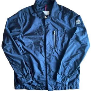 Marinblå vindjacka från Moncler - Snygg marinblå vindjacka från Moncler med klassisk krage, dragkedja framtill och en bröstficka med vit dragkedja. Jackan har Moncler-logga på vänster ärm och justerbara muddar. Perfekt för dig som gillar stilren och sportig look. Storlek 2 vilket motsvarar S/M.