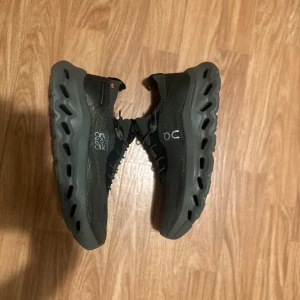 Svarta On Cloudmonster sneakers - Säljer ett par svarta On Cloudmonster sneakers med chunky grå sula och coolt snörningssystem. Skorna har stickad ovandel i syntetmaterial och On-logga på sidan och plösen. Perfekta för dig som gillar sportig och modern stil.