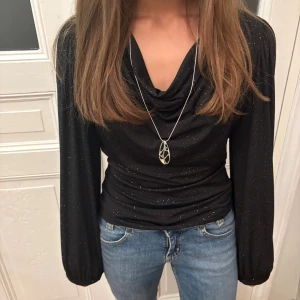 Fest topp  - Snygg svart topp med glittriga detaljer och draperad krage. Långärmad modell med lätt puffade ärmar och figurnära passform. Perfekt för dig som vill sticka ut lite extra. Materialet känns mjukt och följsamt.