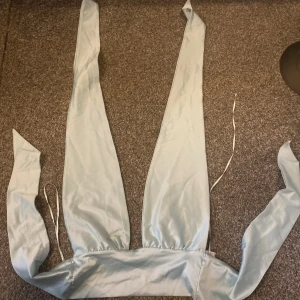 Ljusblå omlottopp i satin - Säljer en ljusblå omlottopp i glansigt satinmaterial. Toppen har långa band som knyts runt kroppen och ger en snygg omlotteffekt. Perfekt för dig som vill ha en trendig och unik look med extra långa knytband.