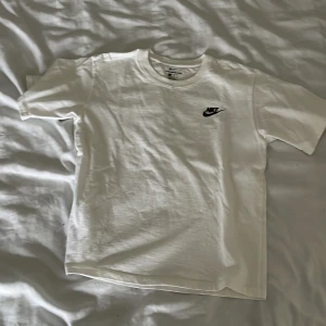 Vit Nike t-shirt i bomull - Säljer en klassisk vit t-shirt från Nike i storlek M. T-shirten har korta ärmar, rund halsringning och en liten svart Nike-logga på bröstet. Tillverkad i mjuk bomull och har en clean, sportig vibe som funkar till allt.