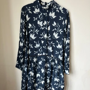 Blommig marinblå klänning från Ganni - Snygg marinblå klänning från Ganni med vitt blommigt mönster. Modellen har hög hals, trekvartsärm och en avslappnad passform med volang nedtill. Materialet känns som mjuk bomull och klänningen har en dragkedja i ryggen för enkel påklädning. The size label was cut off, it suits size 34/36. Never used. 