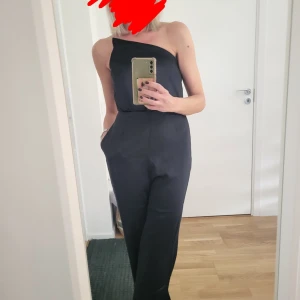 Svart oneshoulder jumpsuit i satin - Supersnygg svart jumpsuit med oneshoulder-design och lång byxdel. Tillverkad i mjukt, glansigt satinmaterial som ger en lyxig känsla. Perfekt för dig som vill sticka ut med en cool och modern look. Sitter skönt med resår i midjan och har fickor på sidorna. Använd en gång.