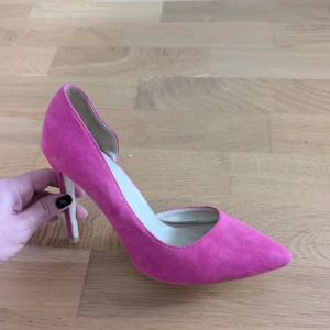 Rosa mocka pumps - Snygga pumps i stark rosa mocka, hög klack. Skorna har en elegant och klassisk siluett som verkligen sticker ut. Perfekta för dig som vill addera färg till din outfit. Insidan är ljus och klacken är smal för en stilren look.