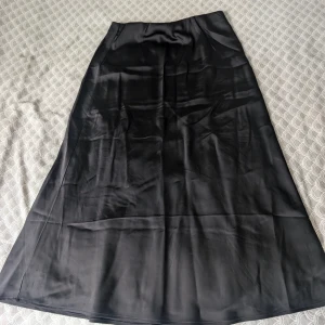 Svart satinkjol, midi - Stilren svart långkjol från HM i glansigt satinmaterial. Kjolen har en klassisk A-linjeform och slät midja för en snygg siluett. Perfekt för dig som gillar en enkel men elegant look.