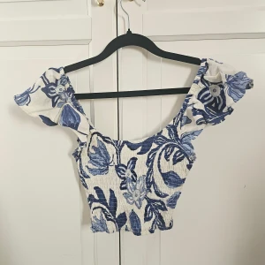 Blommig croppad topp från H&M XS - Superfin croppad topp från H&M i vitt och blått med blommigt mönster. Toppen har volangiga axelband och smockad struktur som ger en snygg passform. Gjord i en skön linneblandning, perfekt för varma dagar. helt ny med ettiket har bara provat