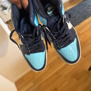 Nike Air Jordan 1 High blå/vit - Säljer ett par Nike Air Jordan 1 High sneakers i blankt skinn med snygg färgkombo av ljusblått, marinblått och vitt. Klassisk siluett med högt skaft, perforerad tå och mörkblå snörning. Perfekta för dig som vill sticka ut med en ikonisk streetstyle.