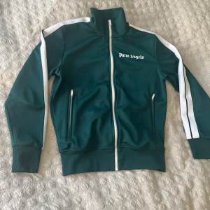 Grön Palm Angels zip-tröja S - Palm Angels zip-tröja i mörkgrönt med vita ränder längs ärmarna och vit dragkedja. Tröjan har hög krage, två fickor med dragkedja och logga på bröstet. Tillverkad i mjuk polyester med ribbade muddar. Perfekt för en sportig och clean look.