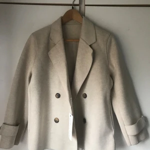 Beige korta kappa med knappar - Snygg beige korta kappamed dubbelknäppning och breda slag. Jackan har långa ärmar med uppvikta muddar och en stilren, klassisk look. Perfekt för dig som gillar minimalistisk stil och neutrala färger.