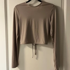 Beige topp öppen rygg från Nelly  - Säljer en oanvänd tröja med snörning i ryggen som går att justera för tajtare eller lösare passform. Passar S och M.