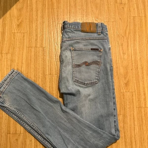Ljusblå Nudie Jeans 32/32 straight fit - Säljer ett par ljusblå jeans från Nudie Jeans i storlek 32/32. Klassisk femficksmodell med orangea sömmar och Nudie-logga på bakfickan. Jeansen har rak passform och är tillverkade i mjukt denimtyg. Perfekt för dig som gillar stilrena och bekväma jeans.