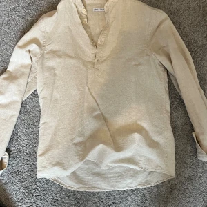Beige henley-skjorta från Zara - Snygg beige henley-skjorta från Zara i mjukt linnematerial. Skjortan har lång ärm, klassisk krage utan slag och knappar vid halsen för en chill och stilren look. Perfekt för dig som gillar enkel och clean stil.