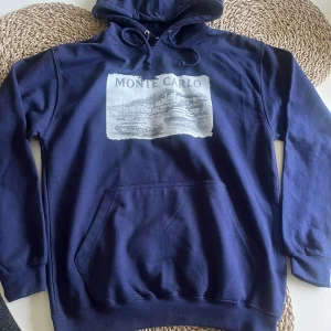 Mörkblå Monte Carlo hoodie med tryck - Snygg mörkblå hoodie med Monte Carlo-tryck på bröstet som visar en klassisk bil och yacht. Tröjan har huva med snörning och en stor magficka. Perfekt för dig som gillar chill och streetwear-vibe. Den är inte använd så helt ny