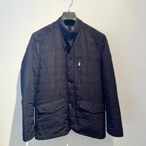 Svart quiltad jacka från Barca - Stilren svart quiltad jacka från Barca med ståkrage och knäppning framtill. Jackan har två stora fickor med lock, en bröstficka med dragkedja och ett snyggt paisleymönstrat innerfoder i blått och beige. Perfekt för dig som gillar klassisk men modern look. Hör av er vid frågor