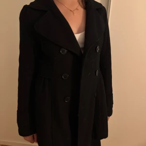 Svart kappa med dubbelknäppning - Stilren svart kappa med bred krage och dubbelknäppning framtill. Jackan har markerad midja och långa ärmar, perfekt för kyligare dagar. Klassisk och tidlös design som passar till det mesta.